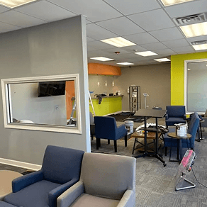 Office lounge with blue chairs, gray walls, and a small table near a bright kitchenette and windowed partition