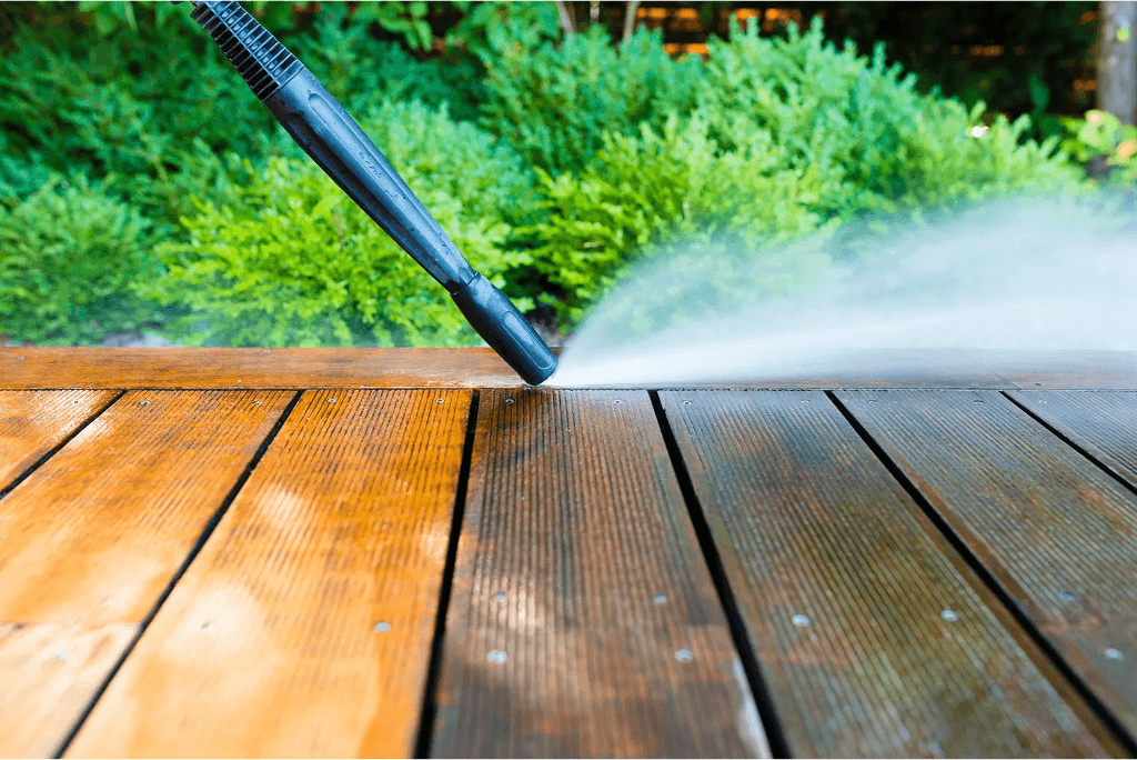 Pressure washer spraying water on a wooden deck outdoors near green shrubs