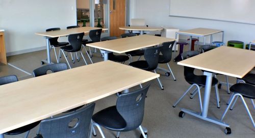 Empty classroom with light wood tables and black chairs.