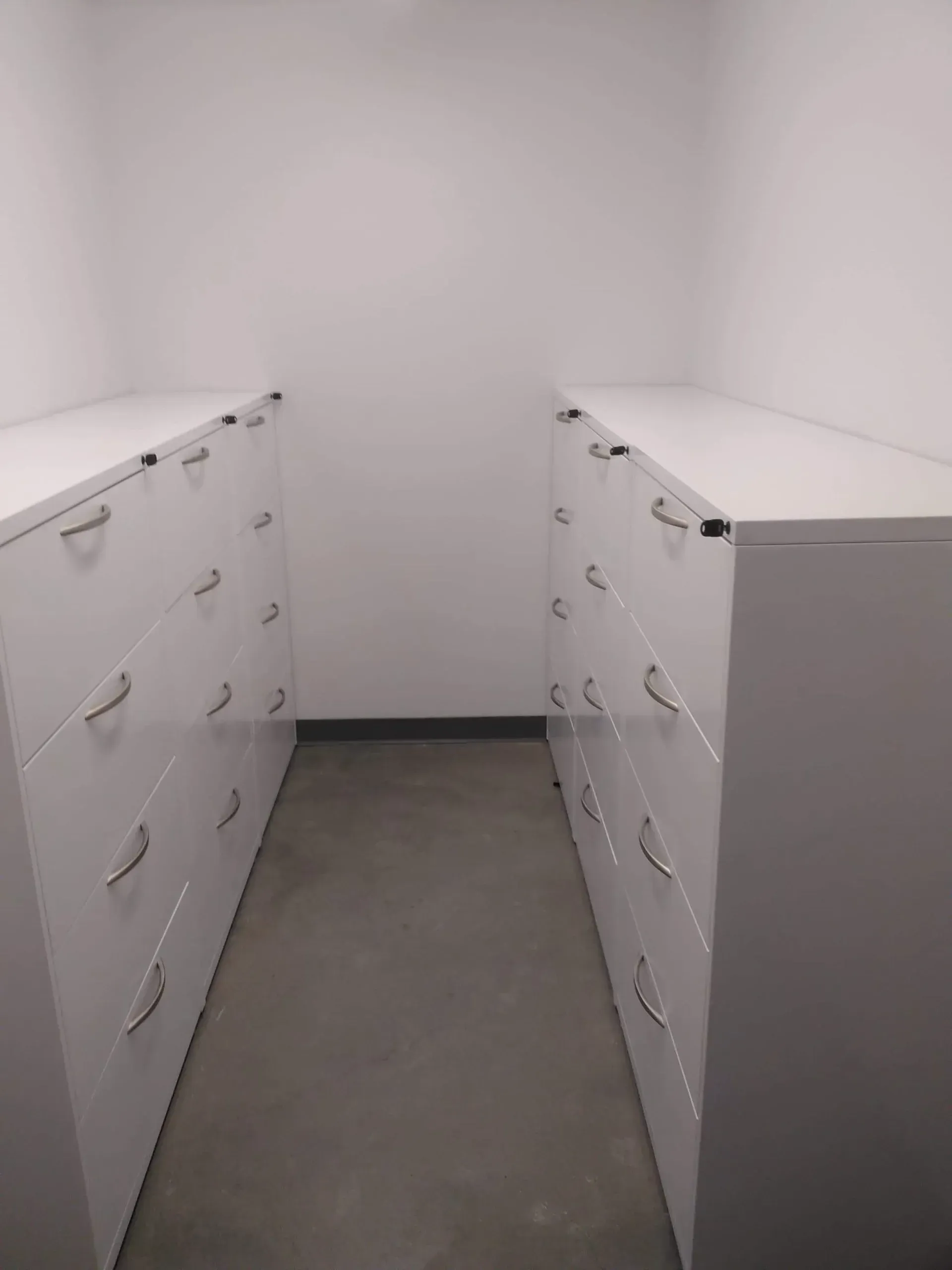 Empty narrow storage room with two rows of white filing cabinets and gray floor.