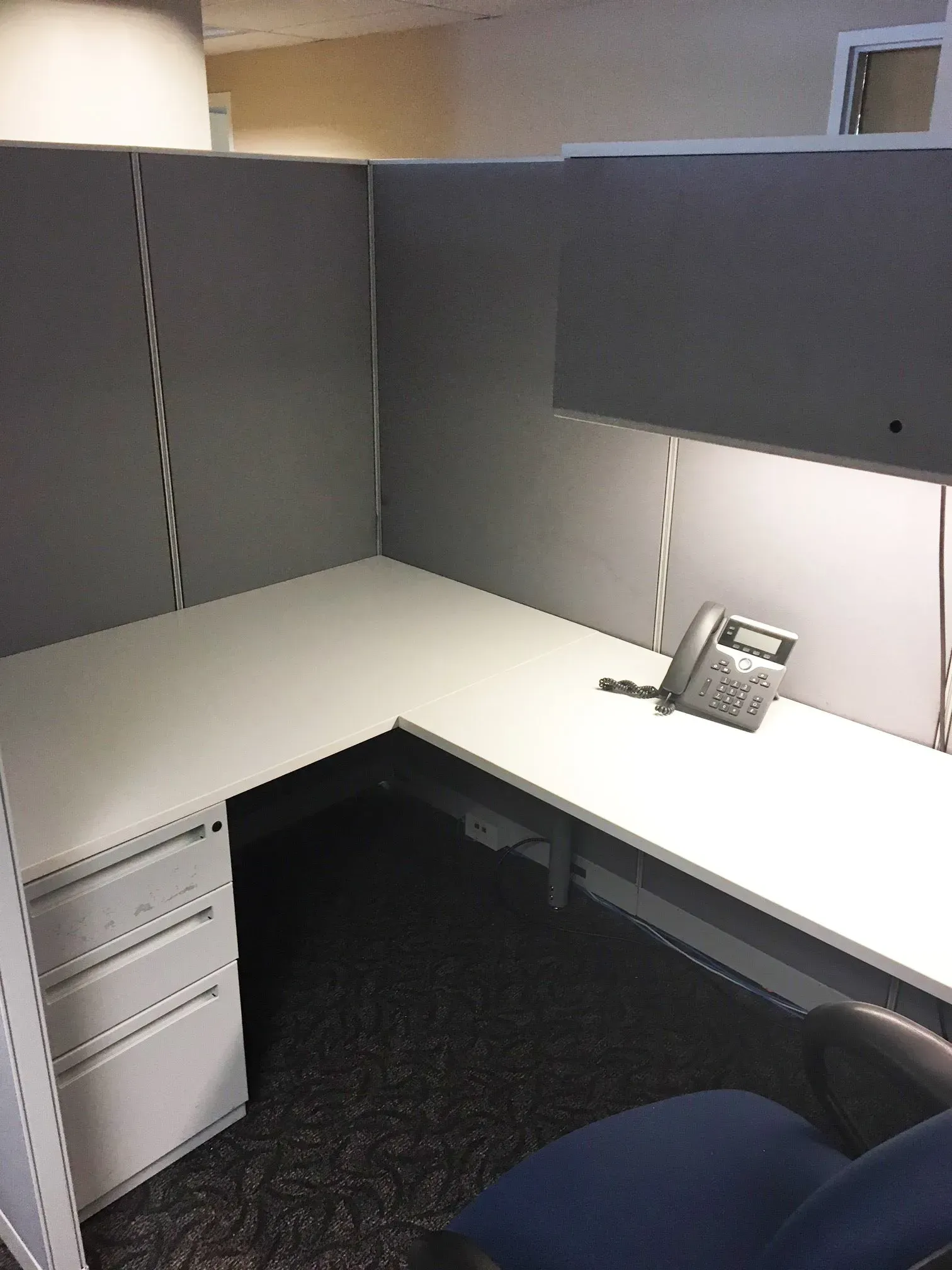 Empty gray office cubicle with desk, phone, drawers, and blue chair in the corner