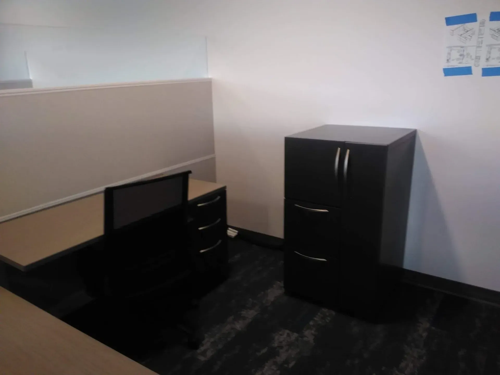 Small office cubicle with black desk, drawer unit, and filing cabinet on a dark carpeted floor.