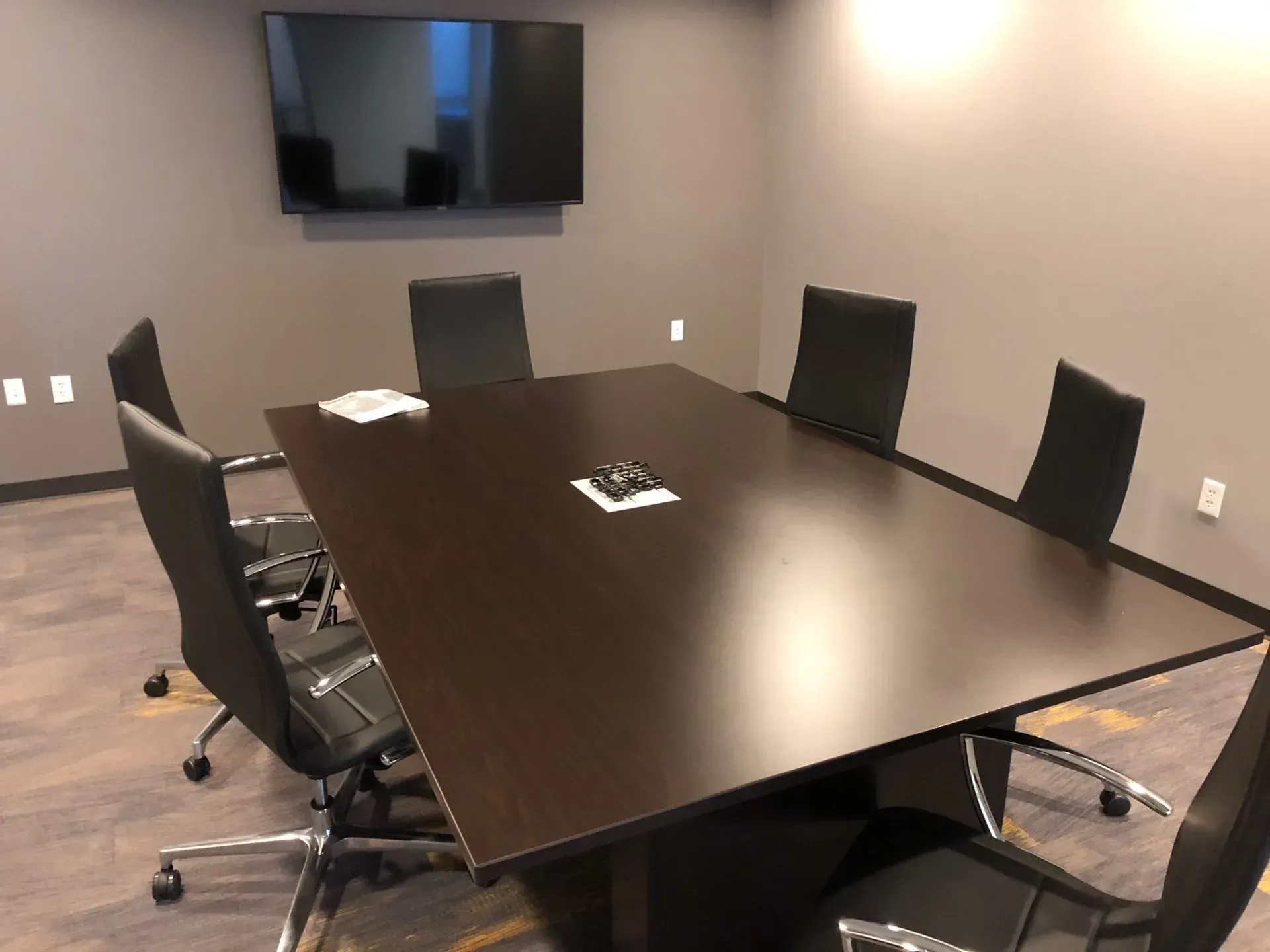 Conference room with dark wood table, black chairs, wall-mounted TV, and light wood flooring.