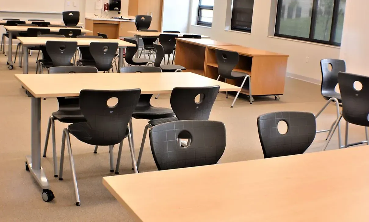 Empty classroom with light wood tables and black chairs arranged in rows.