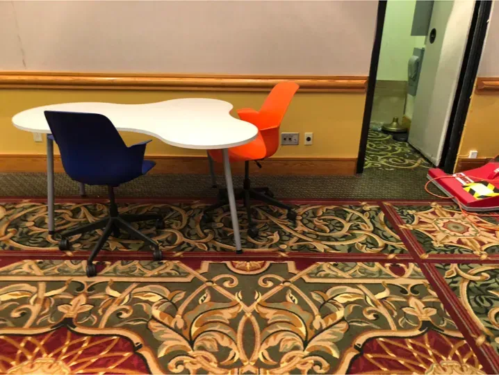 Small white table with blue and orange chairs on patterned carpet in an office lounge.
