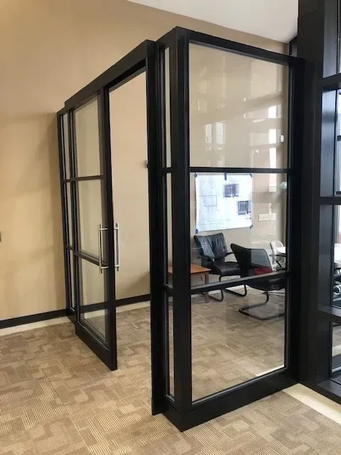 Glass-walled office booth with black frames and a chair inside in a beige hallway