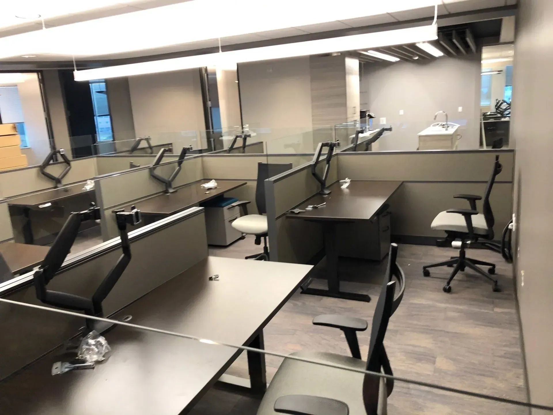 Empty office workspace with rows of desks, monitors, and office chairs under bright overhead lights.