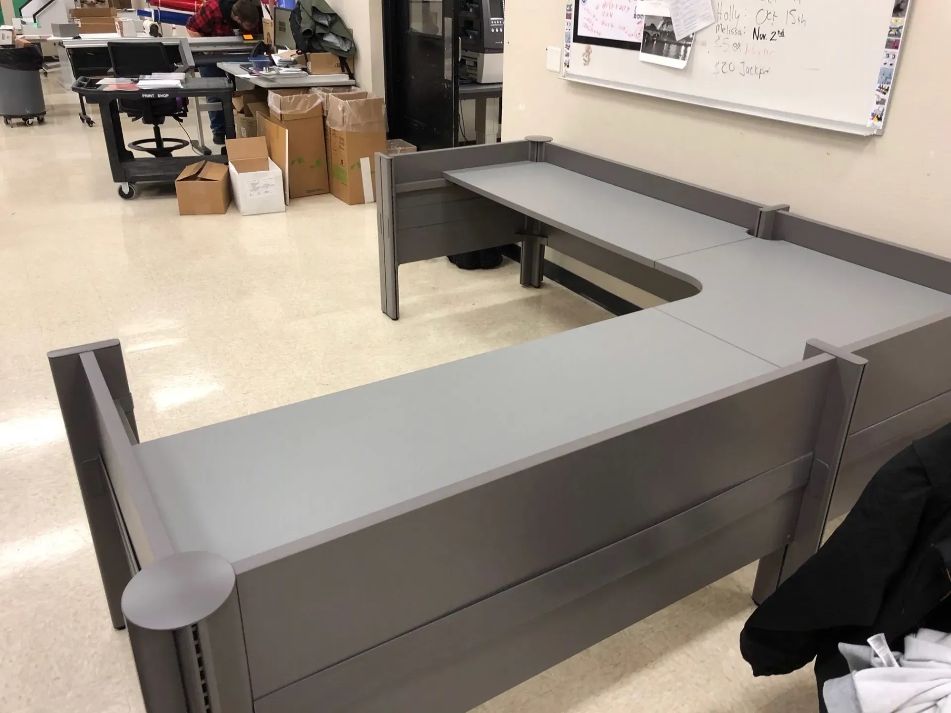 Gray L-shaped office desk in a room with a whiteboard and boxes in the background