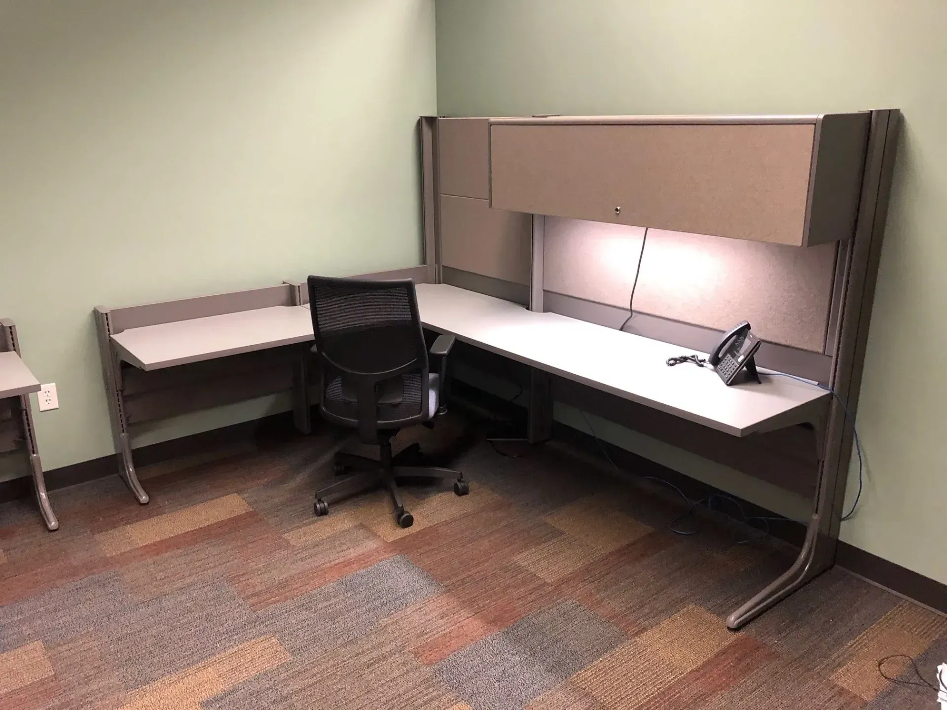 Empty office cubicle with L-shaped desk, black chair, and muted carpeted floor.