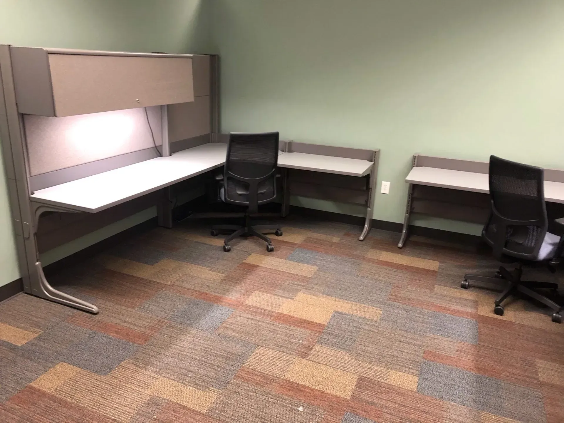 Empty office cubicle with two black chairs, L-shaped desks, and carpeted floor.