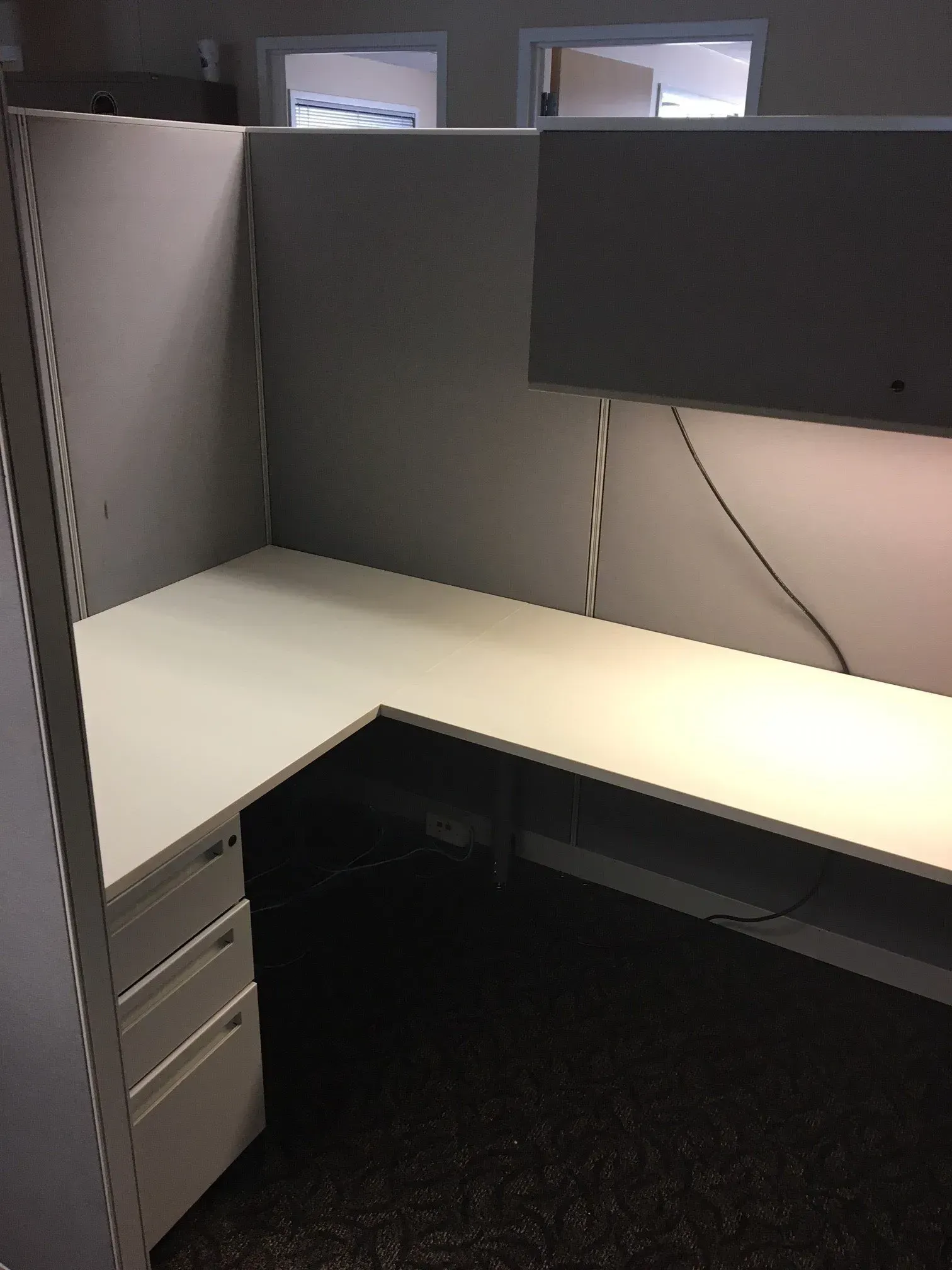 Empty gray office cubicle with a white corner desk, drawers, and wall-mounted cabinet behind it