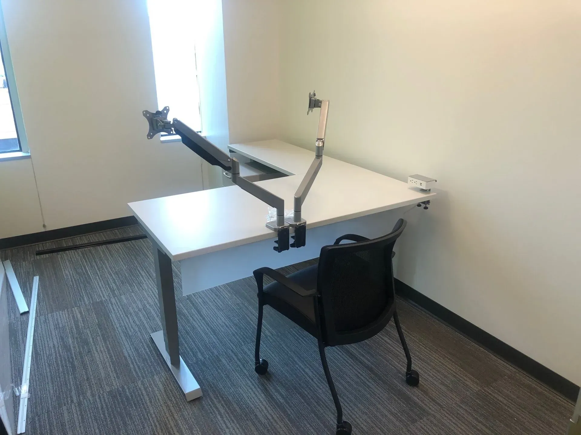Office desk with an articulated monitor arm and a black chair in a sparsely furnished room