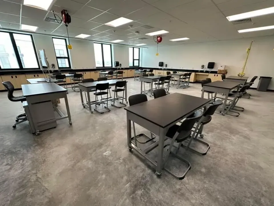 Empty classroom with tables and stools, gray carpet, and large windows letting in daylight.
