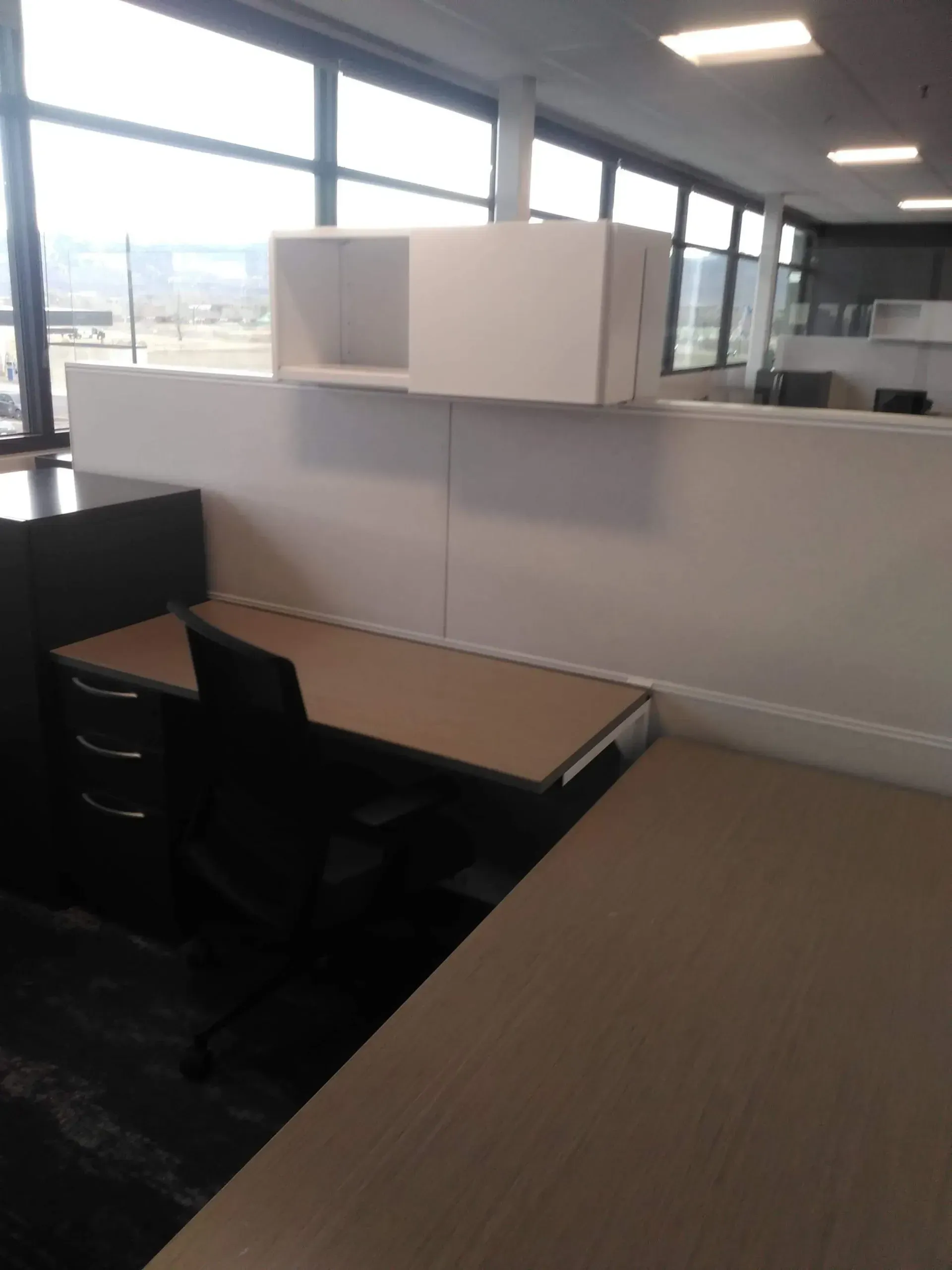 Empty office cubicles with desks, black chairs, and large windows in a bright workspace.