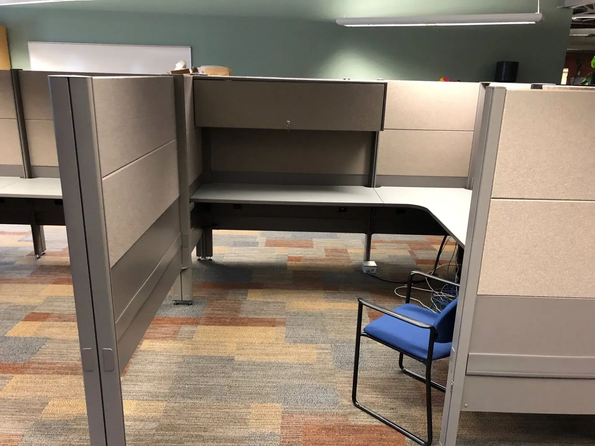 Office cubicle with a black chair and blue cushioned chair under a loft shelf, empty workspace.