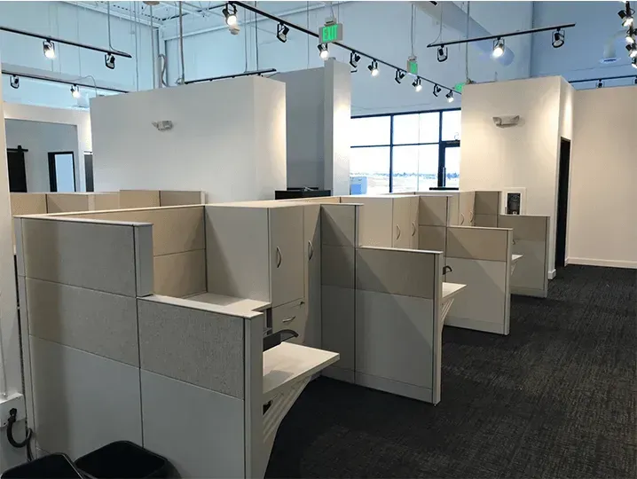 Empty office cubicles in a modern workspace with overhead track lighting and large windows.