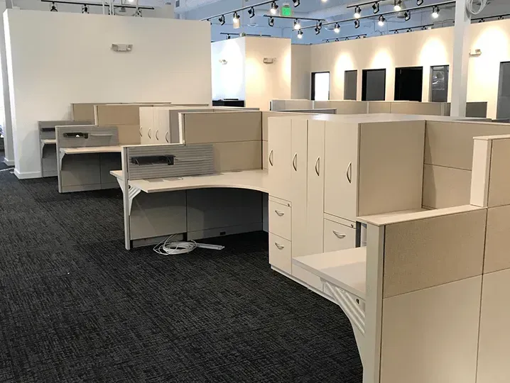 Open-plan office with beige cubicles, cabinets, and workstations on dark carpet.