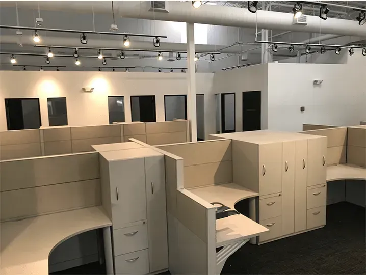 Empty office cubicle workspace with beige partitions, black chairs, and ceiling track lighting