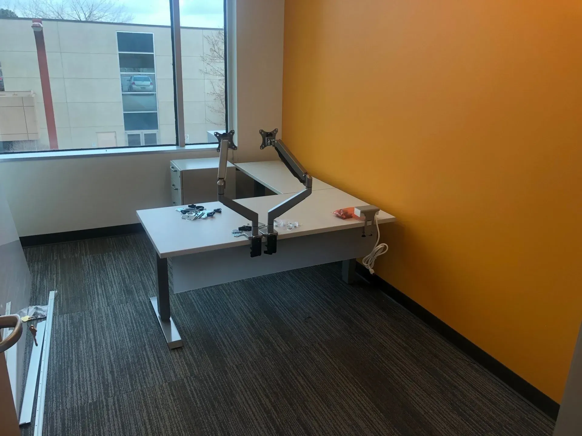 Empty office with a white desk and monitor stand against an orange wall, near a large window.