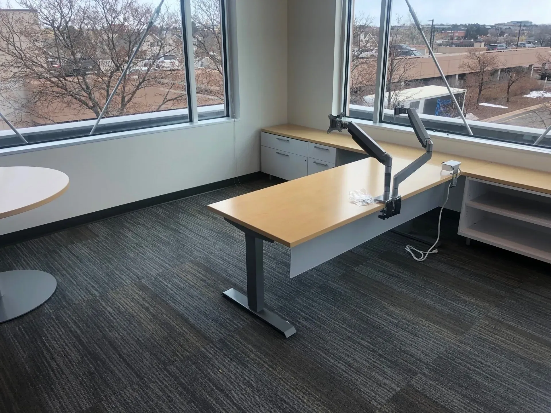 Empty office desk by large windows, with monitor arms and carpeted floor.
