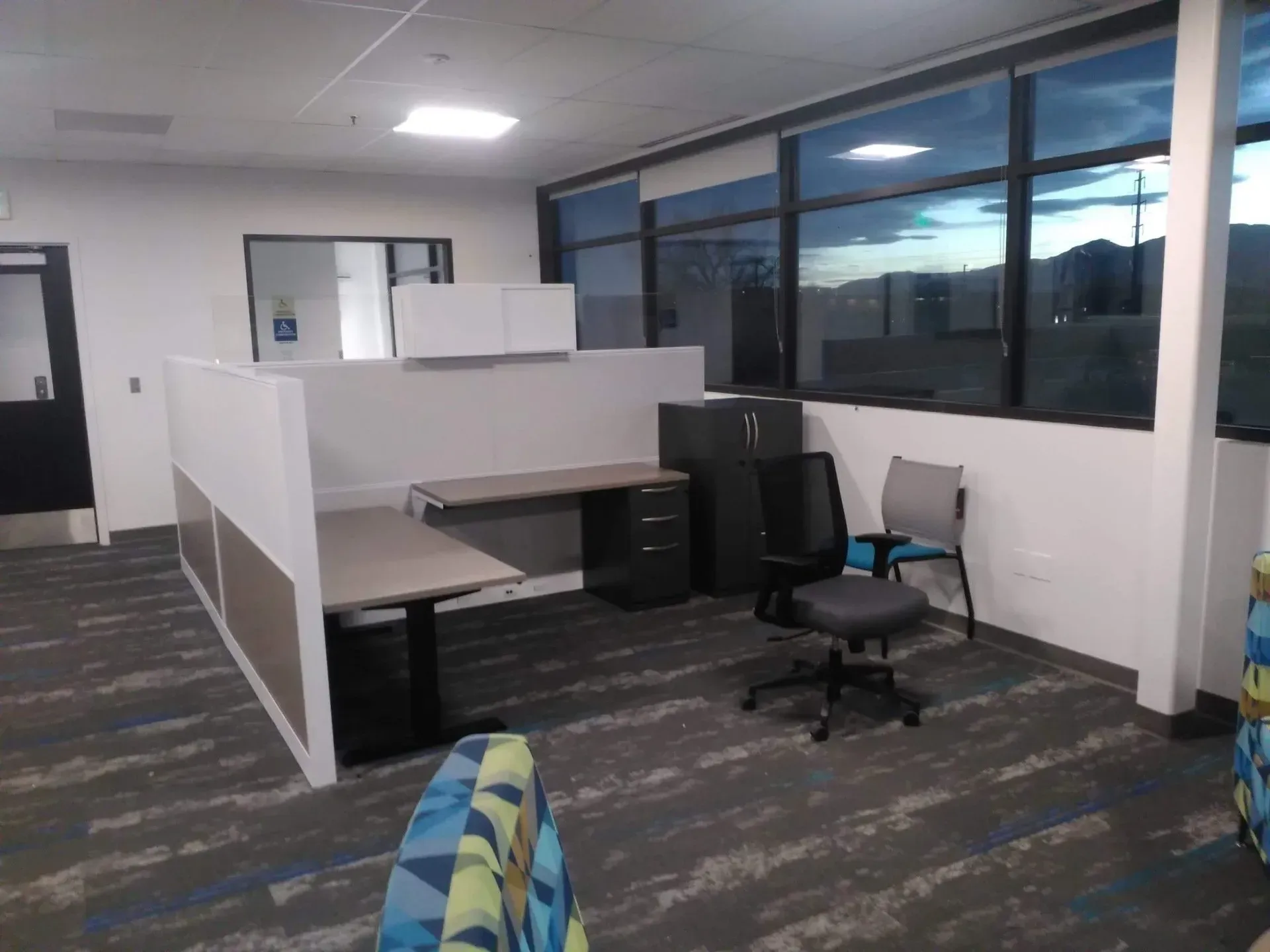 Empty office cubicles with desks, black chair, and large windows overlooking dusk outside.