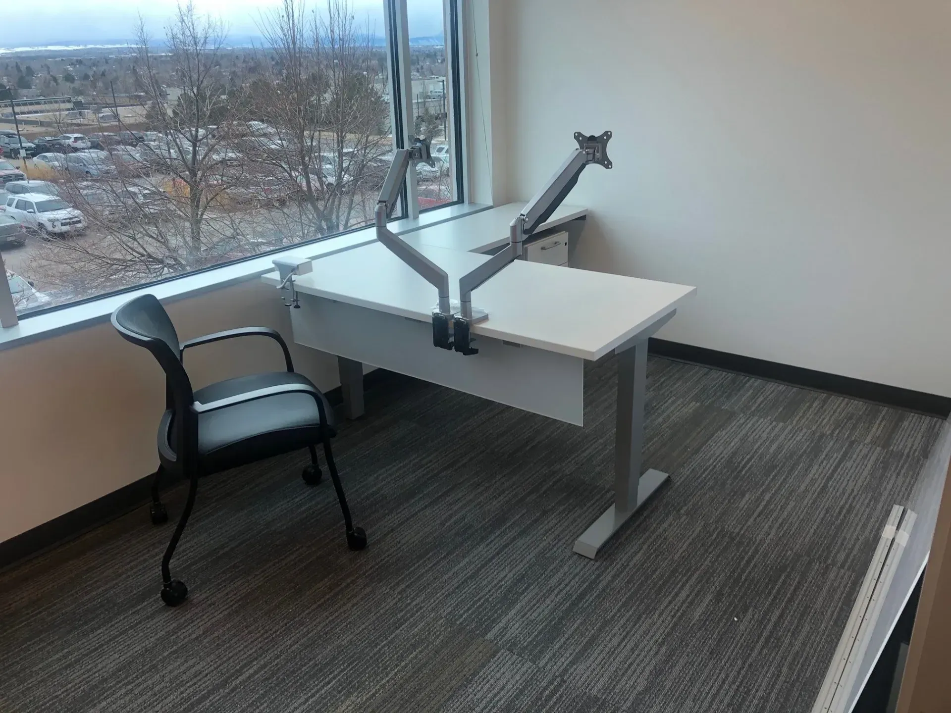 Empty office corner with desk-mounted monitor arm, black chair, and a large window view.