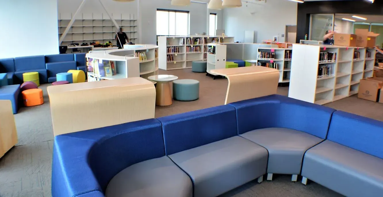 Colorful modern lounge with blue and gray sectional seating, wooden tables, and open shelving in a bright office space
