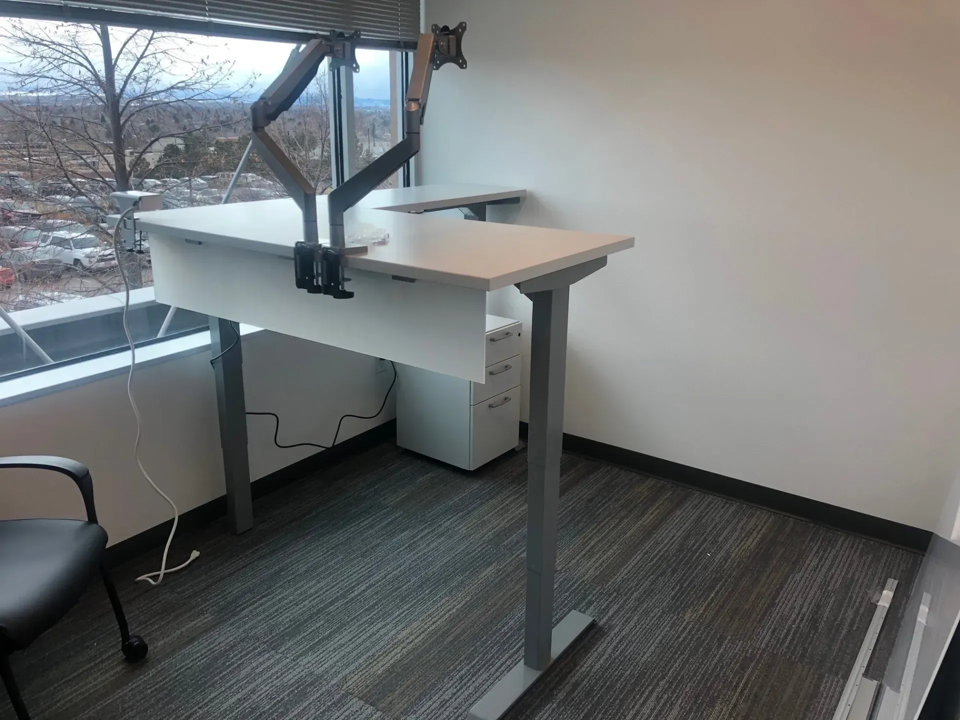 Standing desk by a window in an empty office, with a chair and gray carpet.