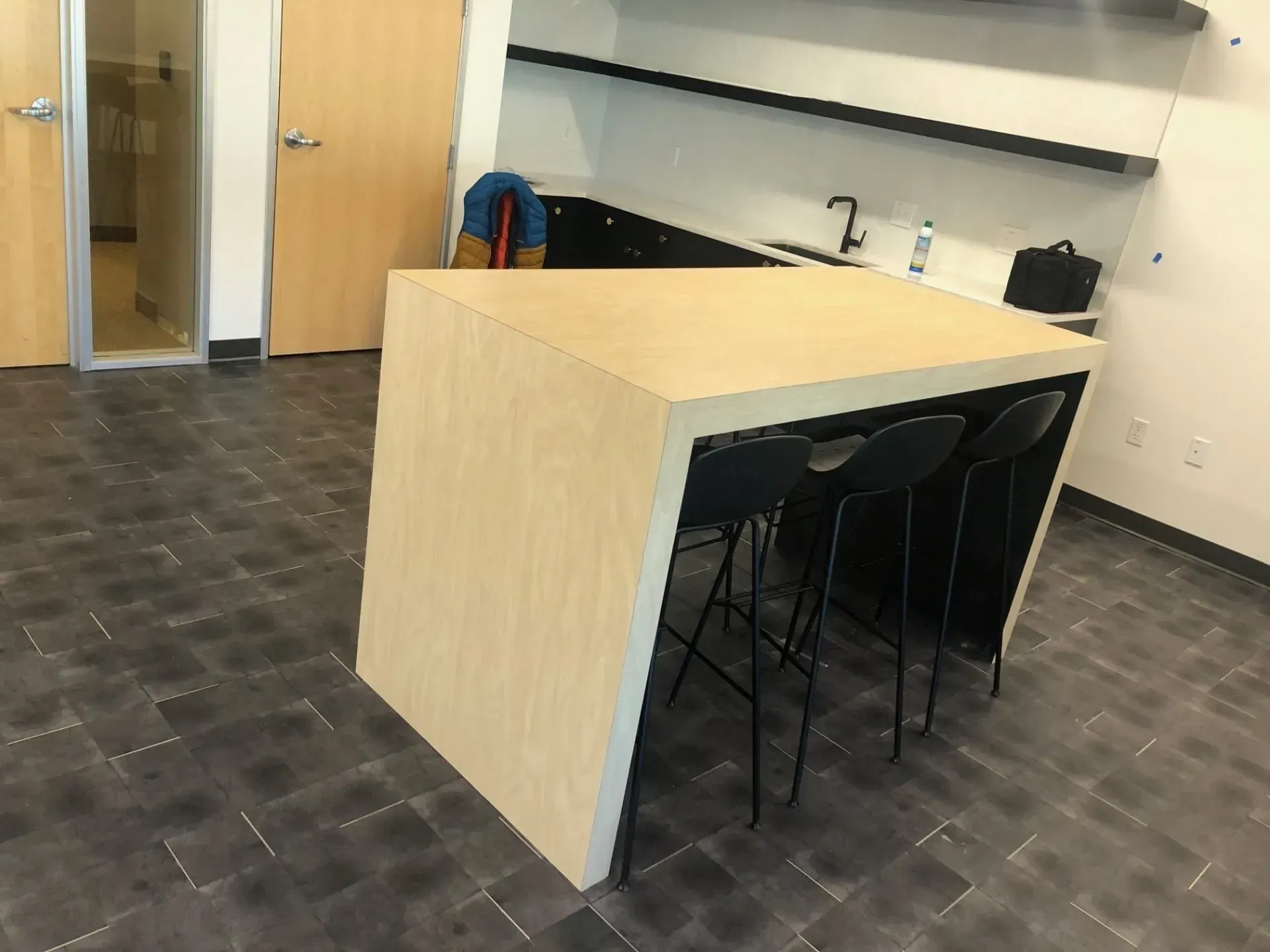 Office kitchenette with a wooden island and black stools beside a sink and shelves