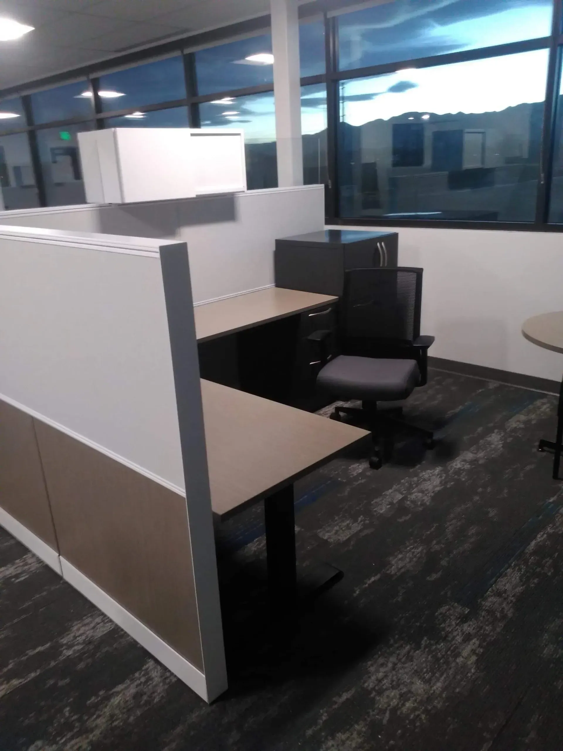 Empty office cubicle with desk, black chair, and large windows at night