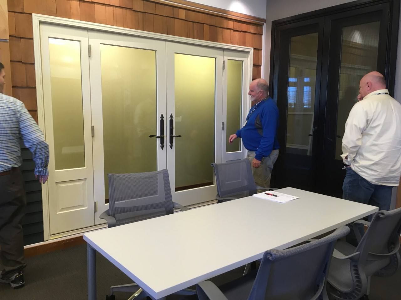 Three men near a conference table in front of glass doors and a wooden wall.