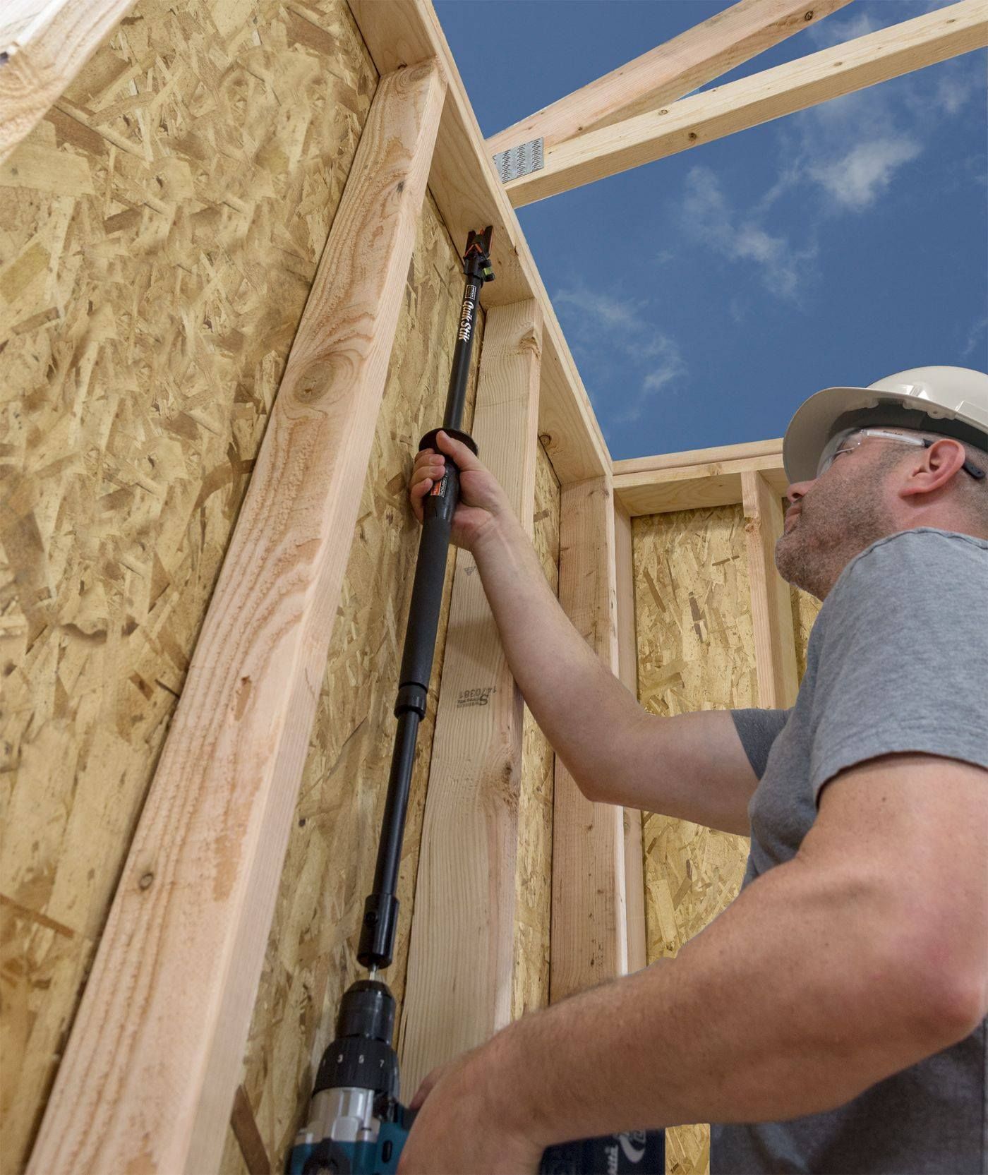 Construction worker using a drill to install a long black tool in a wooden wall under a blue sky.
