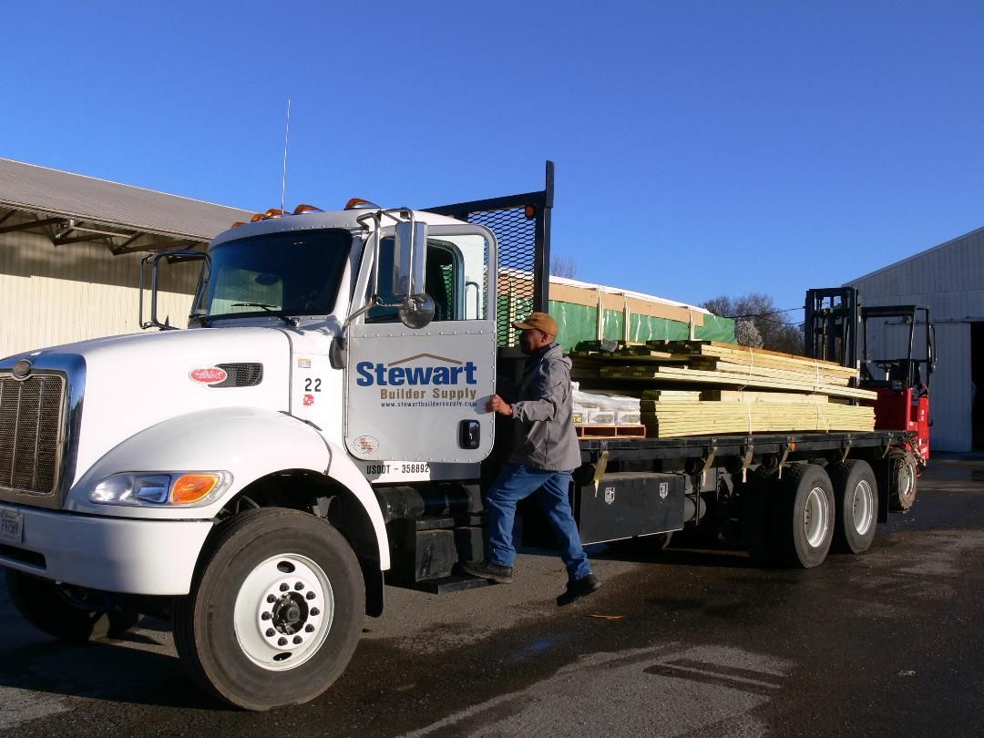 A white flatbed truck with lumber. A person exits the driver's side door. Outdoors, sunny day.