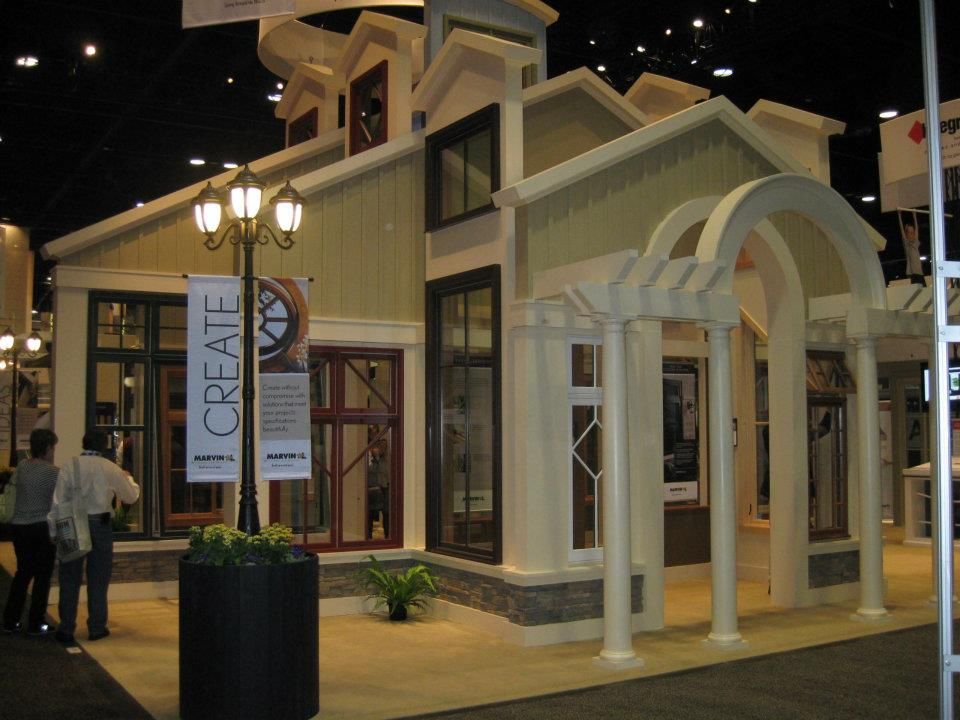 Model house display at a trade show, featuring windows and architectural details. People are nearby.
