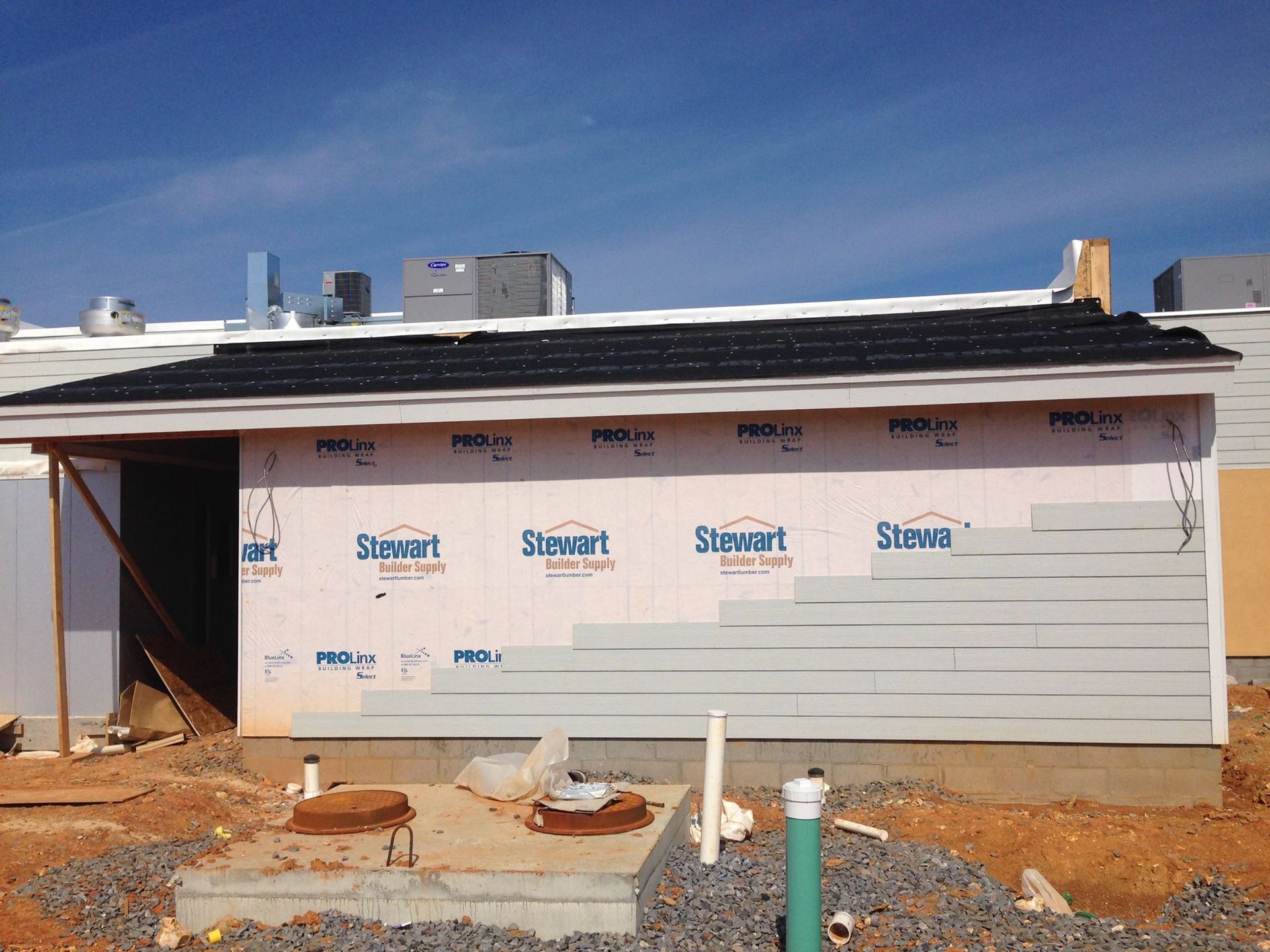 Construction of a building with incomplete siding, gray clapboards, and a black shingled roof under a blue sky.