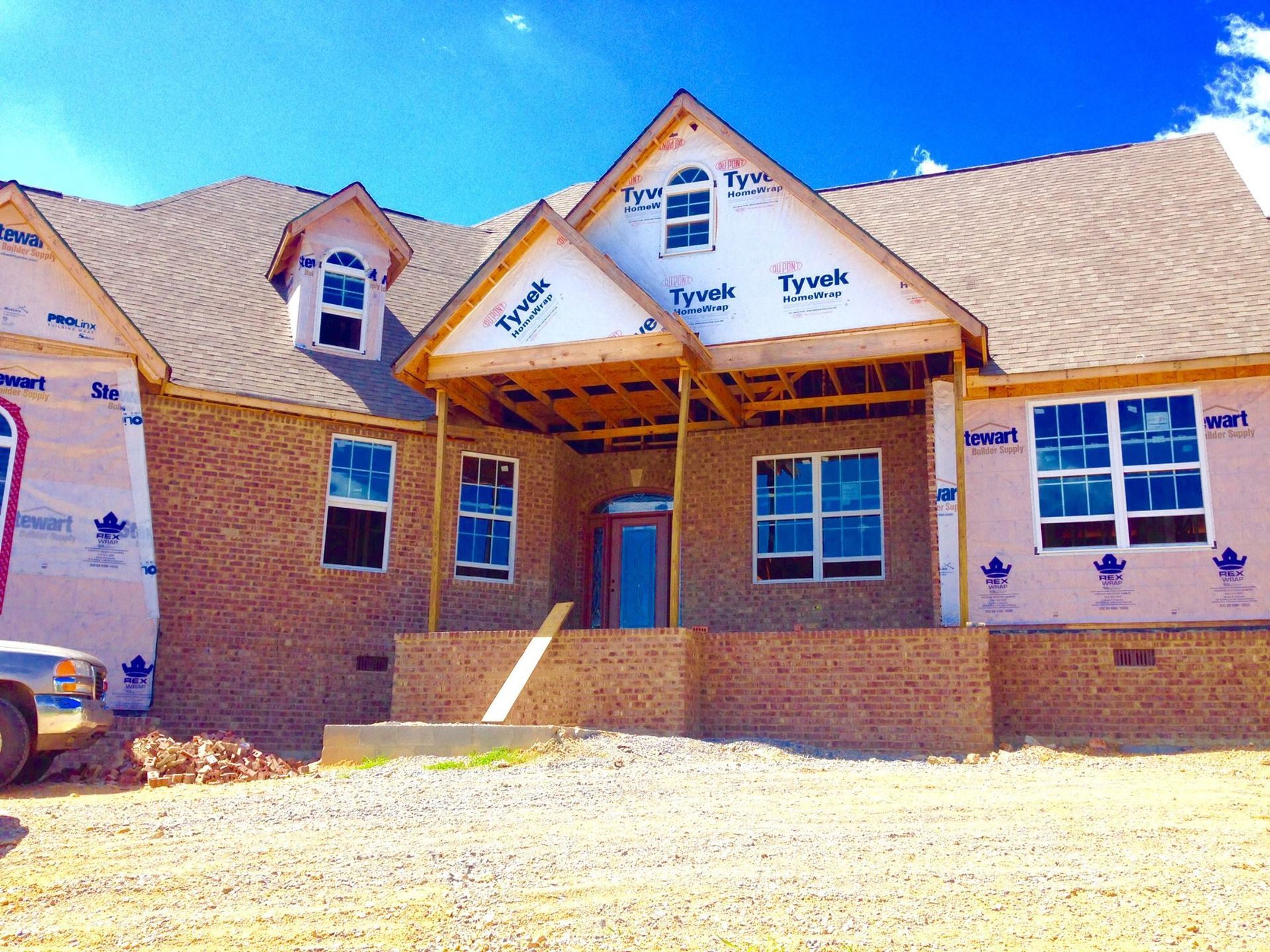 House under construction, brick facade, blue sky, windows, exposed wood beams, tar paper on roof.