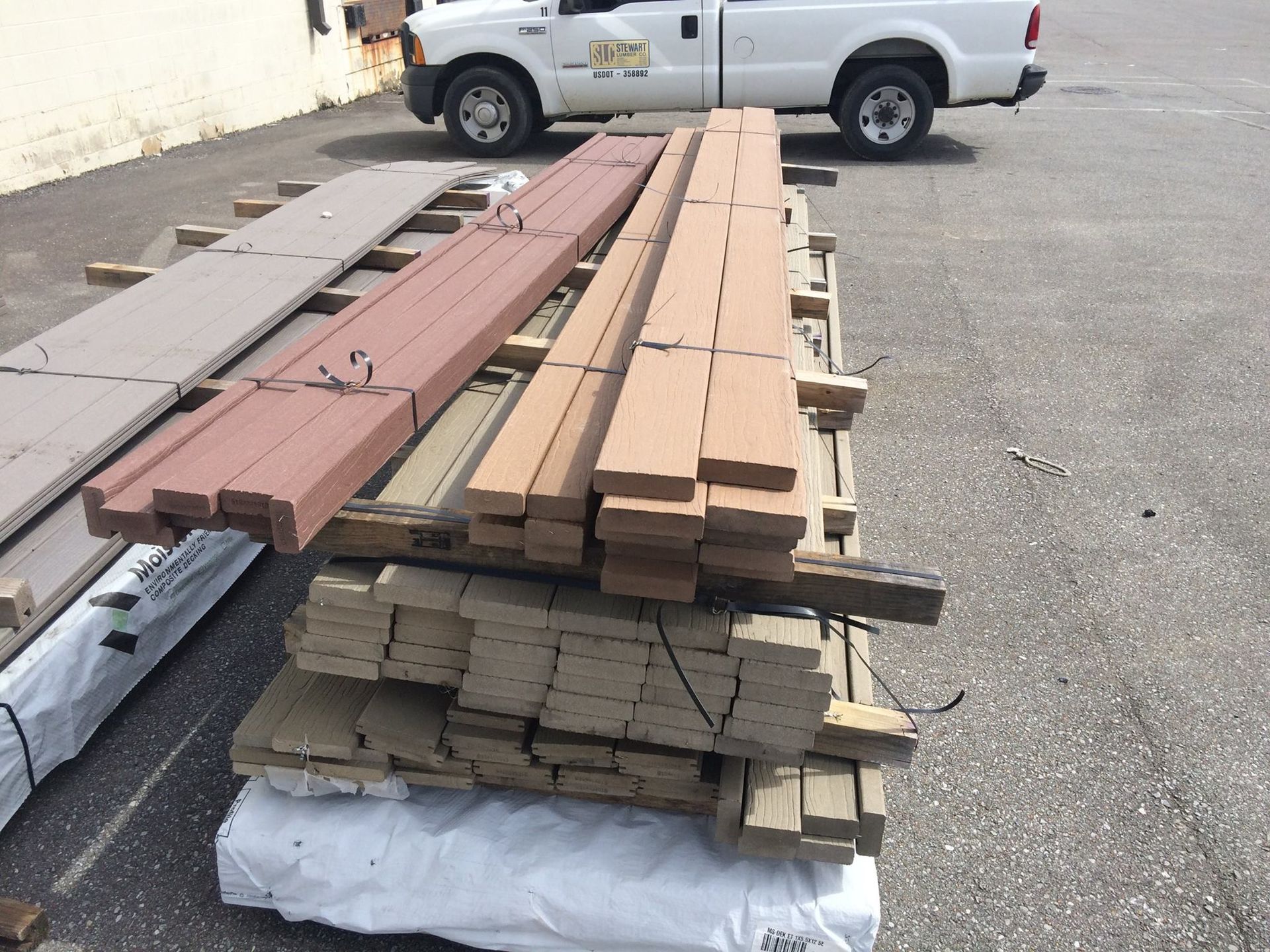 Stacks of lumber in various colors, including brown, red, and gray, on a pallet outside. A white truck is in the background.