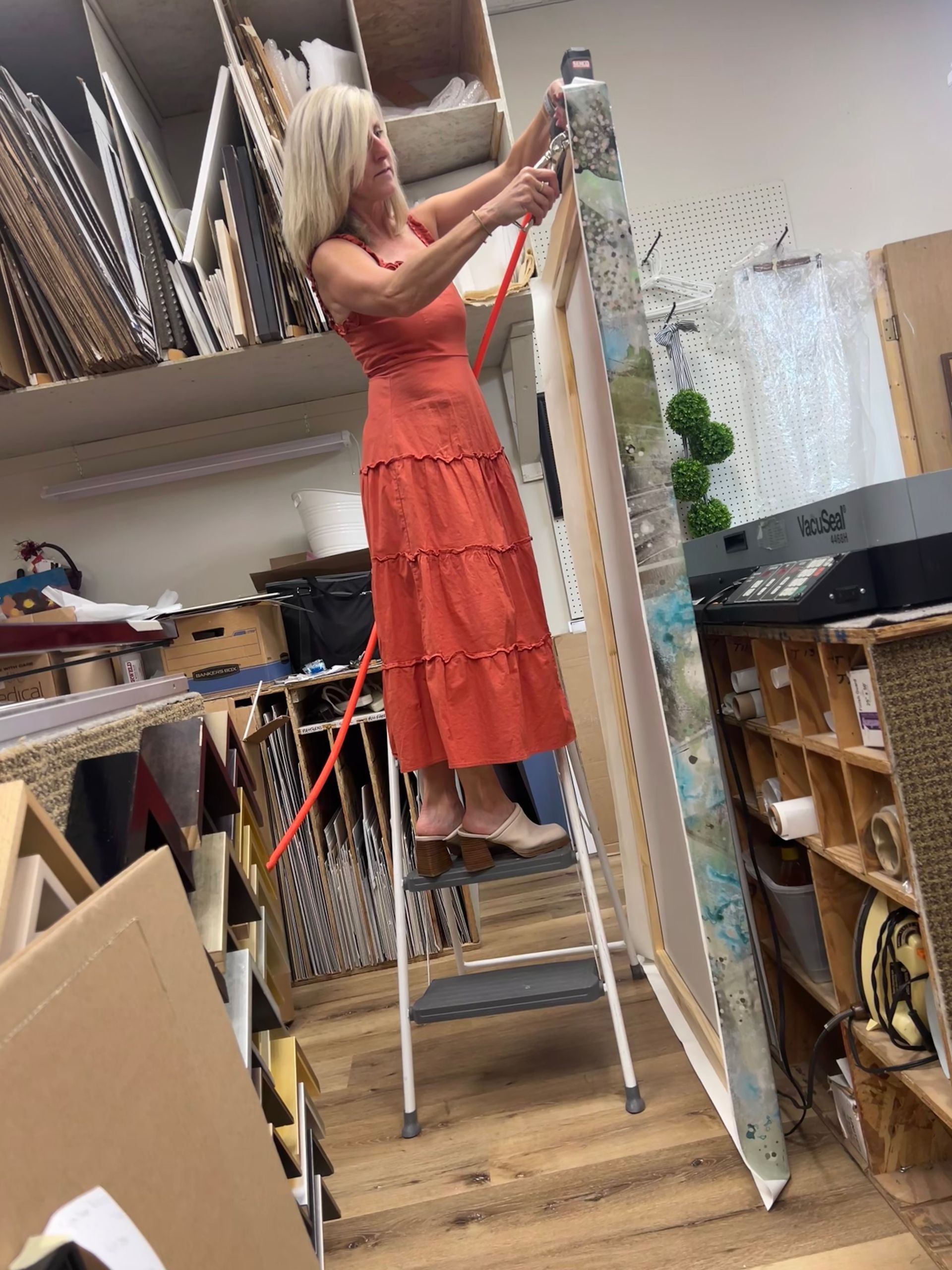 A person in a red tiered dress stands on a step ladder, working on a large framed art piece in a framing workshop.