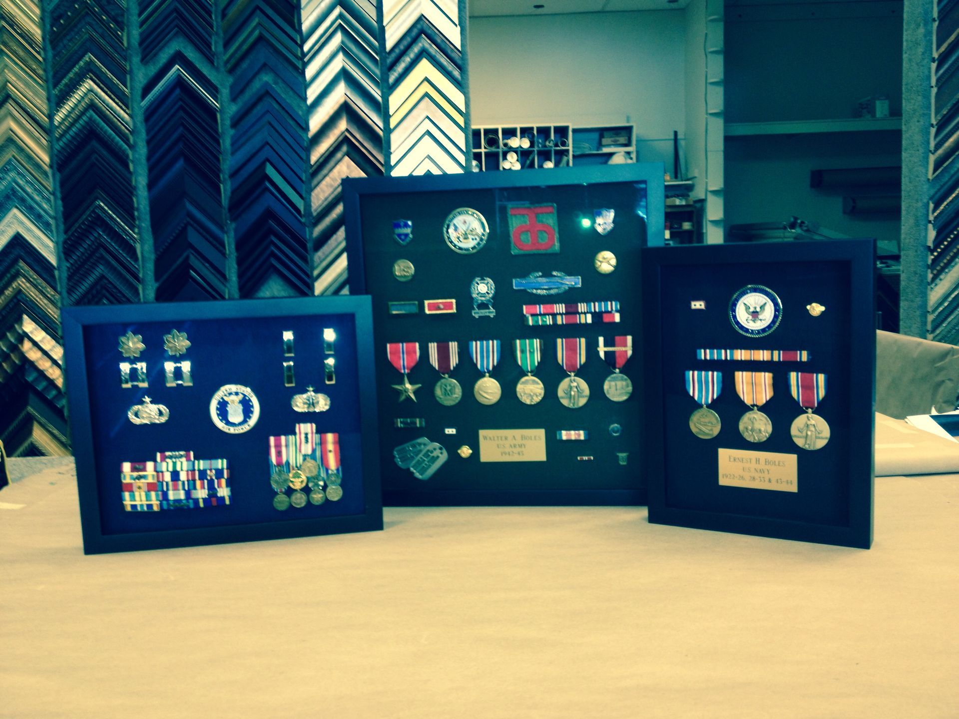 Three shadow box frames filled with military medals and service ribbons, displayed on a table against a wall of frames.