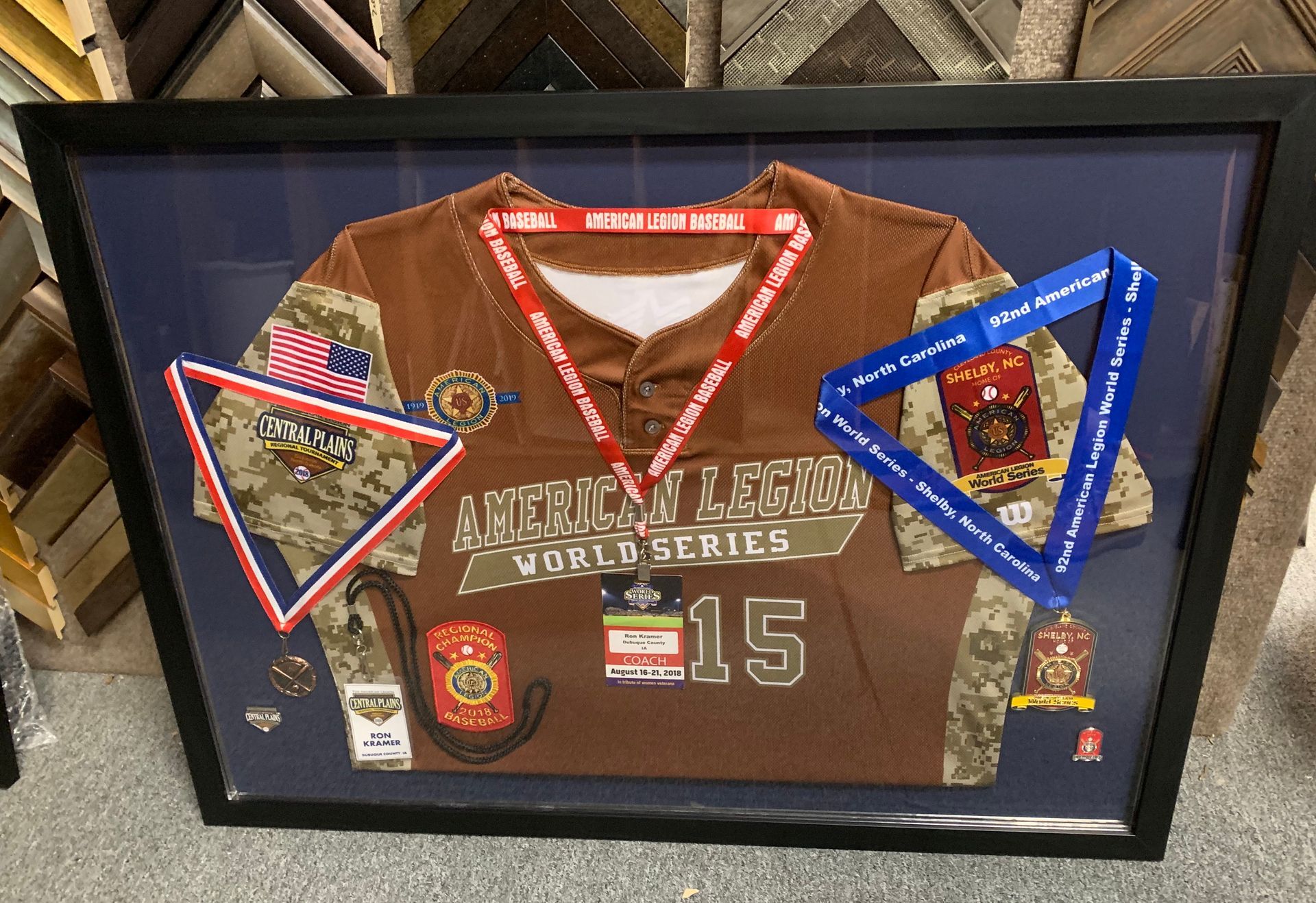 A framed brown American Legion World Series jersey with two medals on ribbons and several commemorative pins displayed.