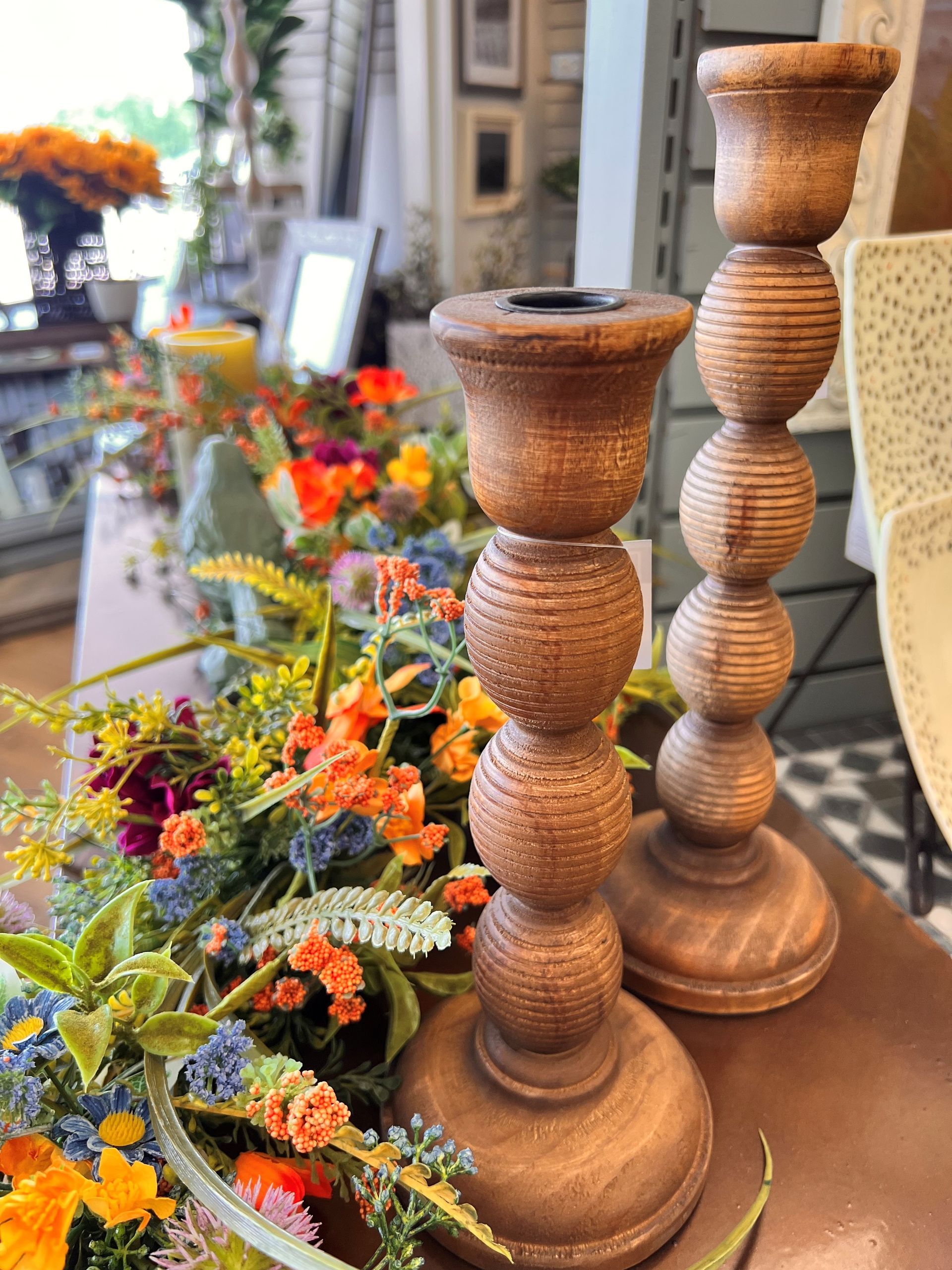 Tall wooden candle holders beside colorful flowers outside a shop