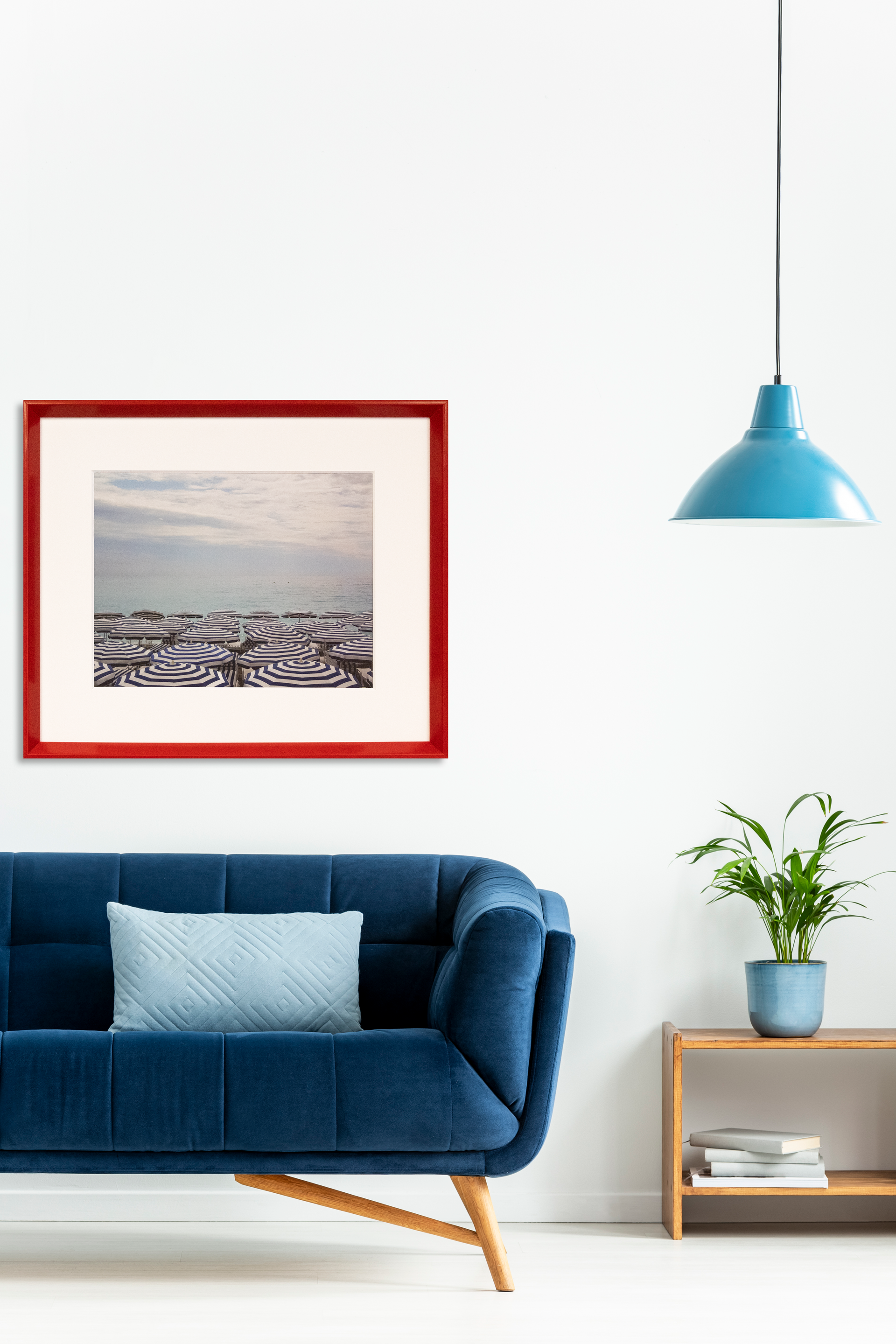 A blue velvet sofa sits next to a side table with a plant, beneath a red-framed print and a blue pendant light.