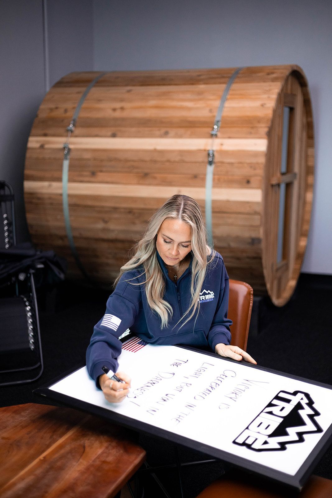 Woman in blue hoodie writing on a whiteboard, wooden barrel sauna in background.