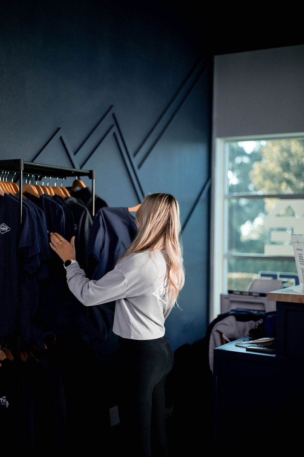Woman browsing clothing rack in a store, dark blue wall with geometric design.