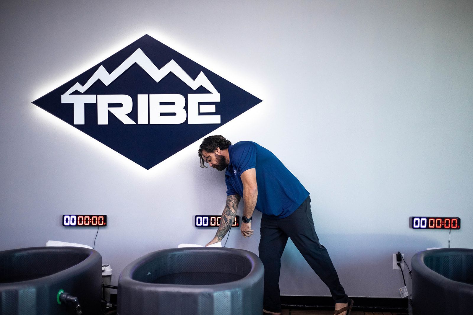 Man in a blue shirt cleaning a cold plunge tub at Tribe gym, with sign and timers.