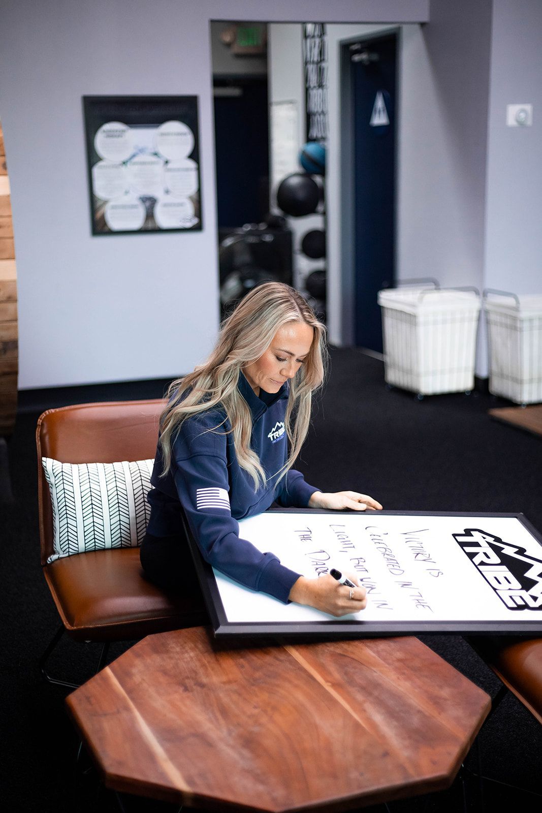 Woman with long blonde hair writing on a whiteboard, seated on a brown leather couch in a modern setting.