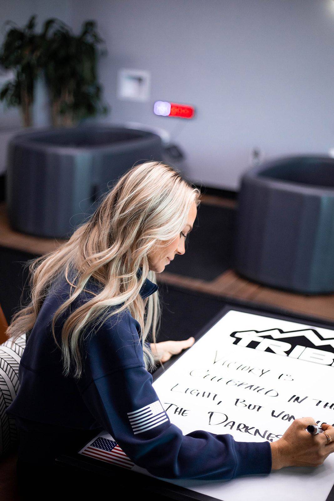Woman writing on a large white surface; interior setting with dark sweater and two dark round objects.