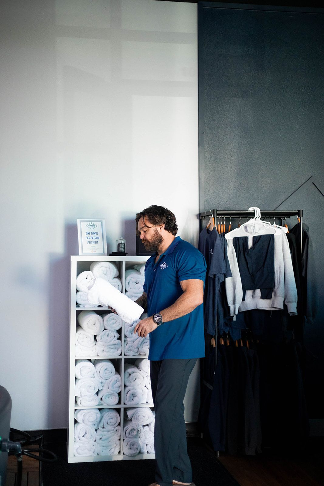 Man in blue shirt, gray pants, looking at towels in a white shelf, next to clothes rack.