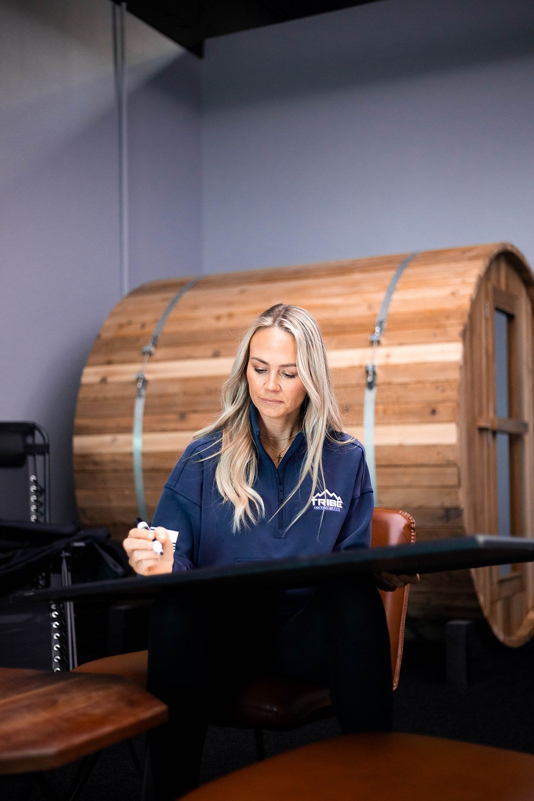 Woman in blue top seated, writing, with wooden sauna barrel in background.