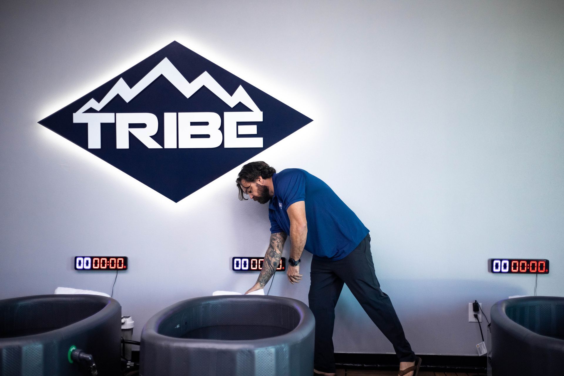 Man preparing ice bath at a "Tribe" wellness center. Dark tubs, white logo, digital timers.